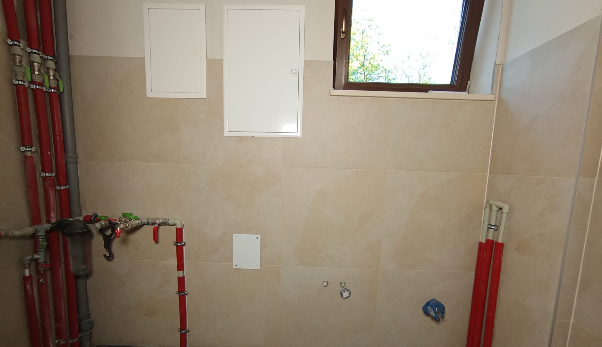 Beżowe płytki na ścianie w łazience z widocznymi rurami grzewczymi w kolorze czerwonym i białymi drzwiczkami rewizyjnymi oraz oknem.
