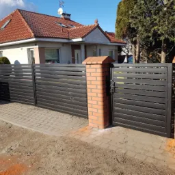 Nowoczesna, pozioma brama wjazdowa z metalu w kolorze antracytu, osadzona na ceglanych słupkach, na tle domu z czerwoną dachówką i błękitnego nieba.