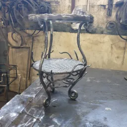 Metalowy kwietnik z dwoma półkami, wykonany ręcznie z surowego metalu, widoczny w warsztacie ślusarskim.