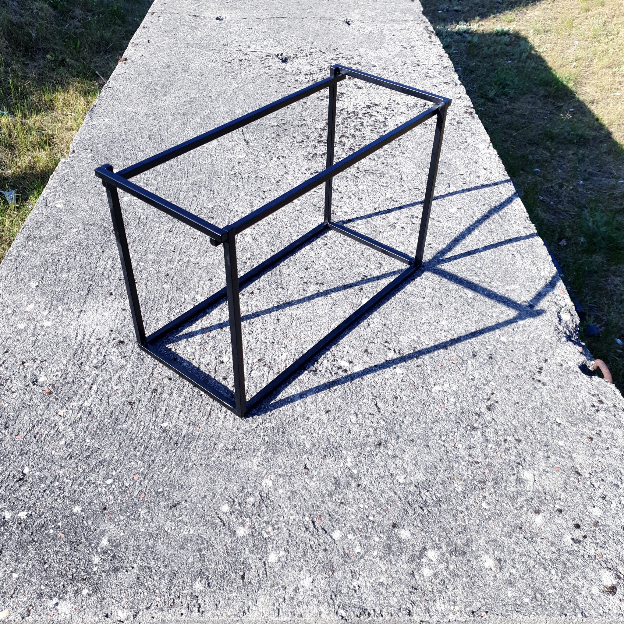 Surowy, czarny szkielet metalowej konstrukcji, prawdopodobnie elementu balustrady lub schodów, ustawiony na szarej betonowej powierzchni w słoneczny dzień.