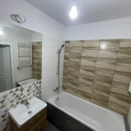 Nowoczesna łazienka z wanną w zabudowie, umywalką na szafce z drewnopodobnym dekorem i lustrem, akcentem są trójkątne płytki dekoracyjne przy umywalce oraz płytki imitujące drewno na ścianie przy...