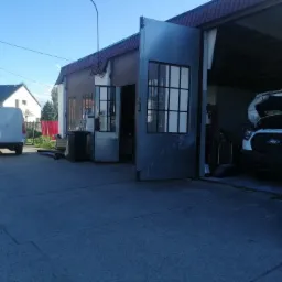 Warsztat samochodowy z otwartymi wrotami, biały van z podniesioną maską wewnątrz, drugi biały van zaparkowany na zewnątrz, budynek warsztatu z metalowymi drzwiami i oknami.