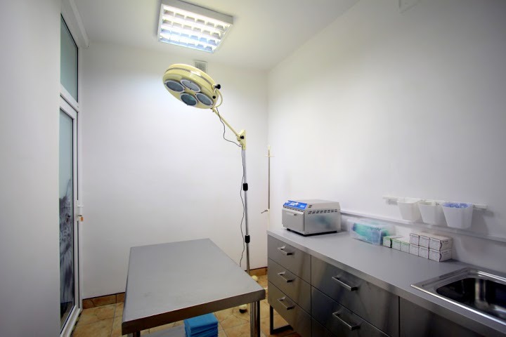 Jasne, minimalistyczne wnętrze gabinetu zabiegowego z metalowym stołem i lampą operacyjną na statywie; na blacie ze stali nierdzewnej sterylizator i pojemniki z akcesoriami.