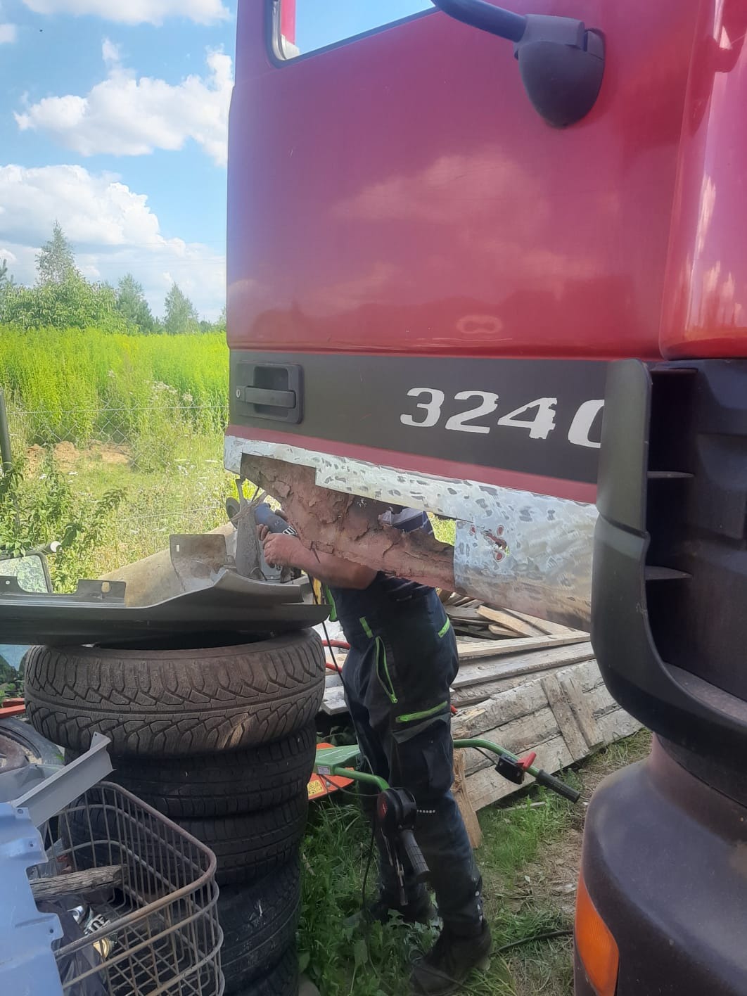 Pracownik w kombinezonie szlifuje uszkodzony fragment karoserii czerwonej ciężarówki, model 3240, na zewnątrz, w tle zieleń i niebo.