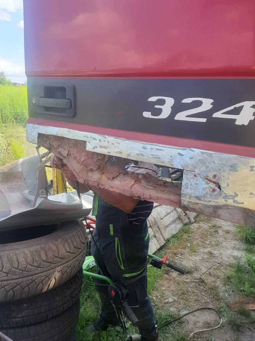 Uszkodzony bok czerwonej ciężarówki z widoczną korozją i śladami napraw blacharskich, pracownik w kombinezonie roboczym w trakcie pracy, stos opon w tle.