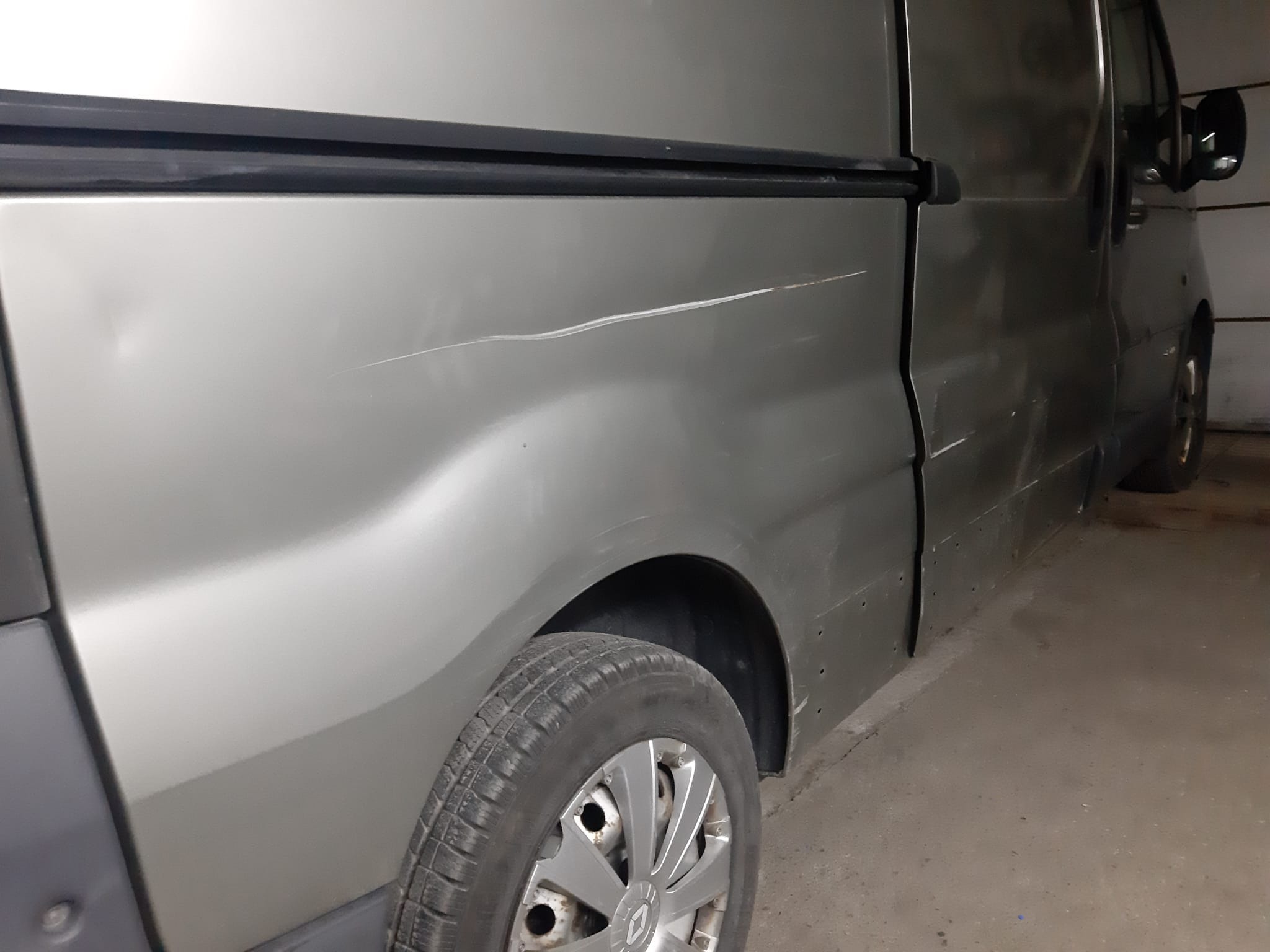 Bok srebrnego vana z wyraźną rysą na karoserii i widocznymi śladami korozji, zaparkowanego w słabo oświetlonym garażu.
