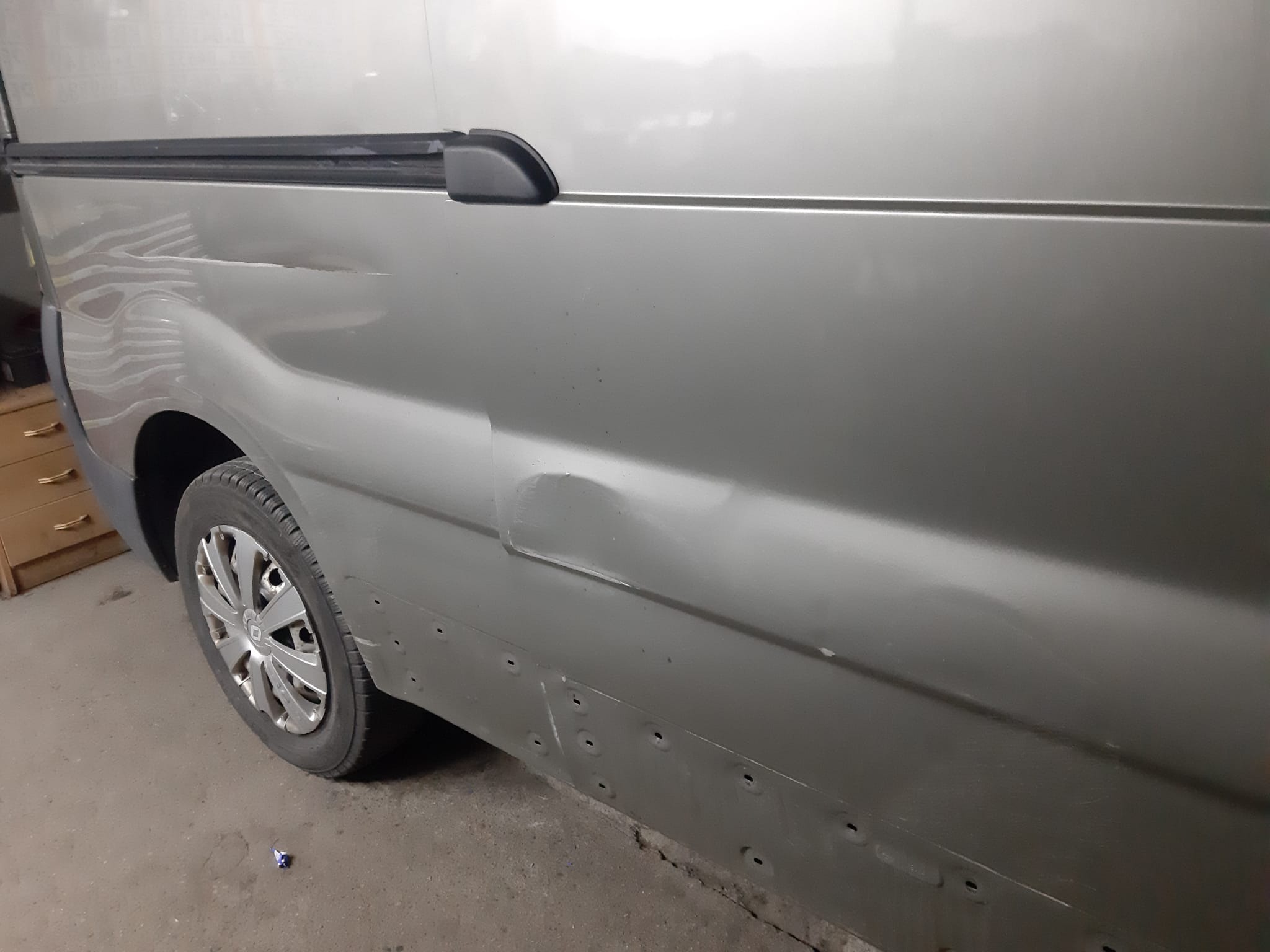 Uszkodzony bok szarego samochodu dostawczego z widocznym wgnieceniem i śladami po spawaniu punktowym przygotowanego do naprawy blacharskiej.