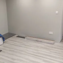 Jasne panele podłogowe ułożone w pustym pokoju z szarymi ścianami, widoczna paczka paneli Kronostep Flooring przy ścianie.