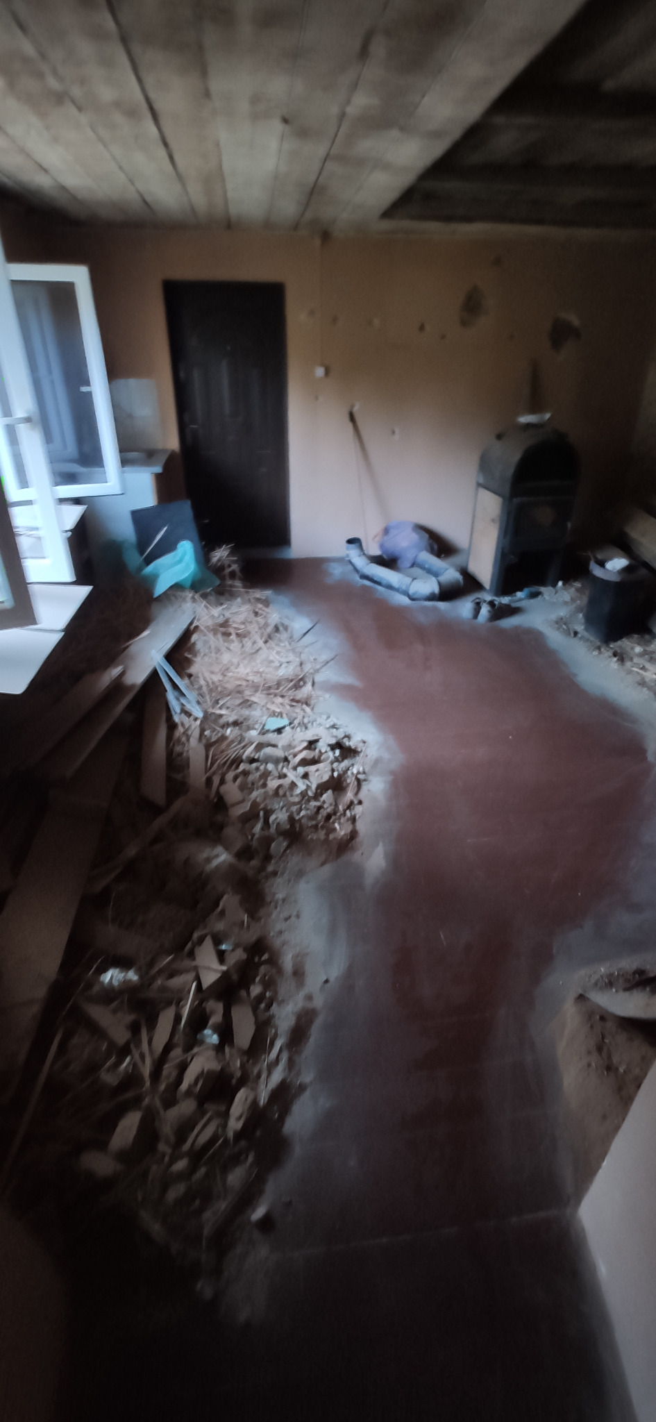 Wnętrze pokoju w trakcie remontu z widocznymi resztkami płytek, gruzem, nowymi oknami i starym piecem.