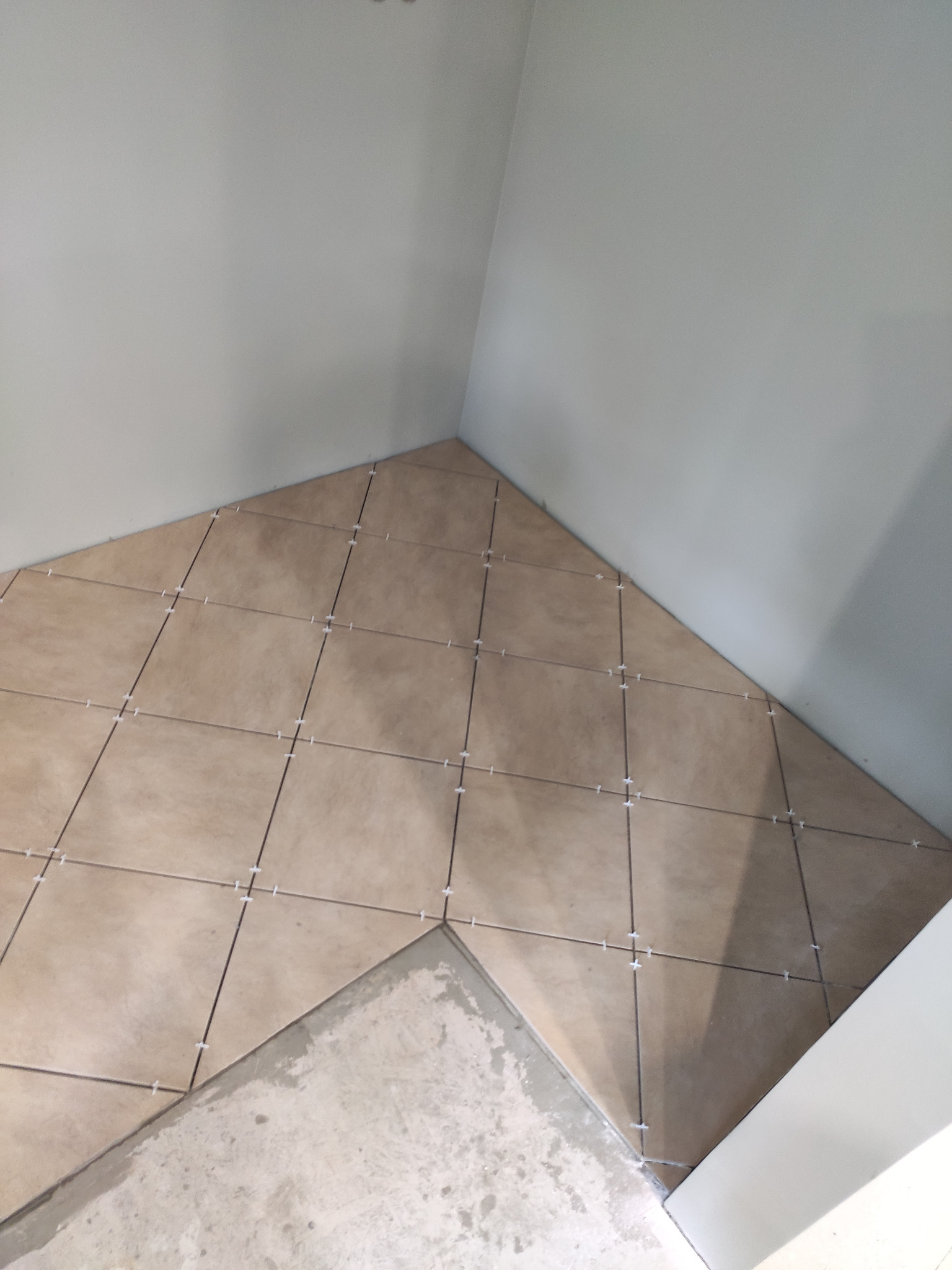Układanie beżowych płytek podłogowych w rogu pomieszczenia, z widocznymi krzyżykami dystansowymi i fragmentem surowej wylewki.