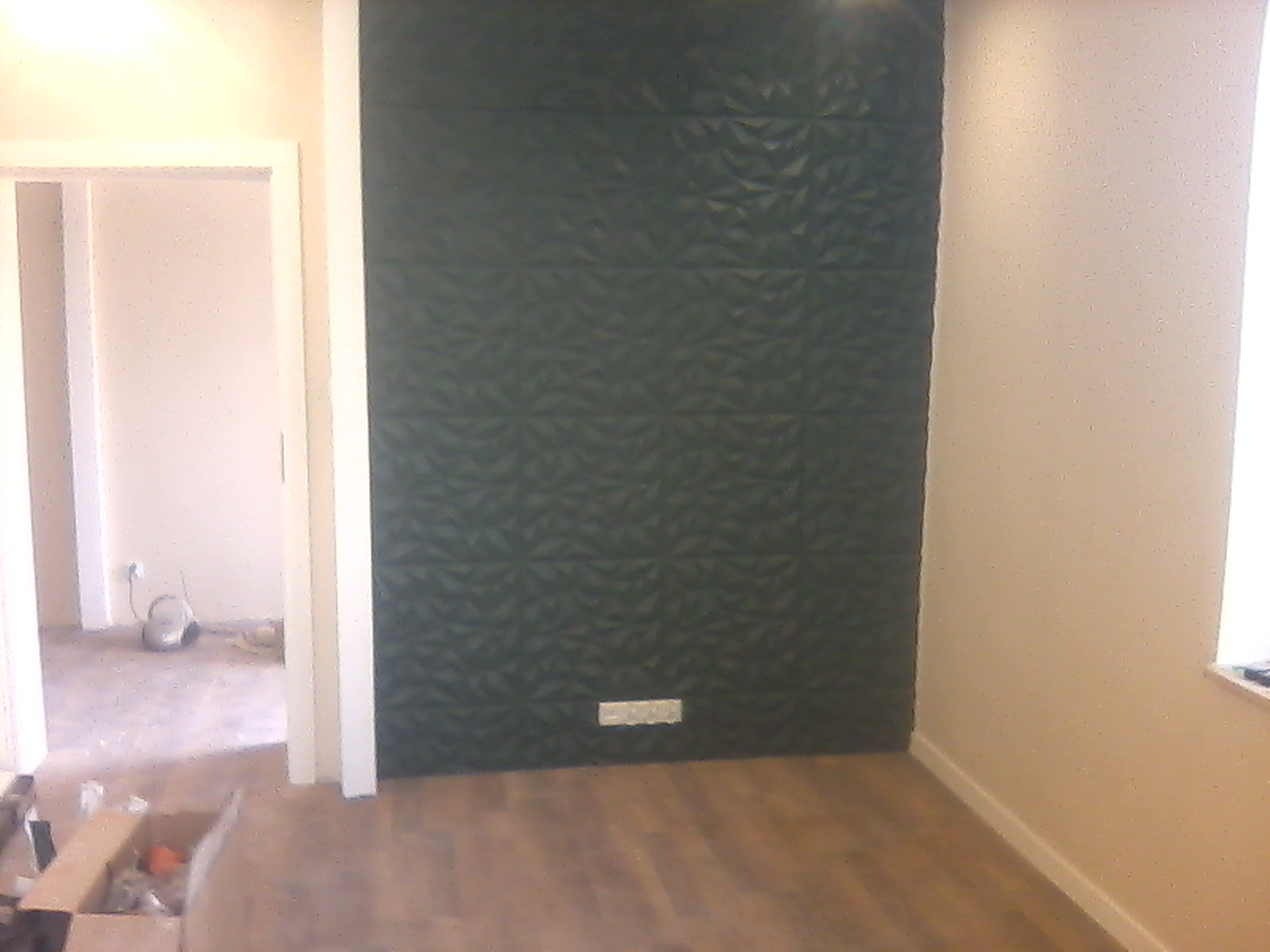 Wykończona ściana z ciemnozielonych paneli 3D o wzorze geometrycznym, widoczna w nowym pomieszczeniu z drewnianą podłogą i gniazdkiem elektrycznym na dole ściany.