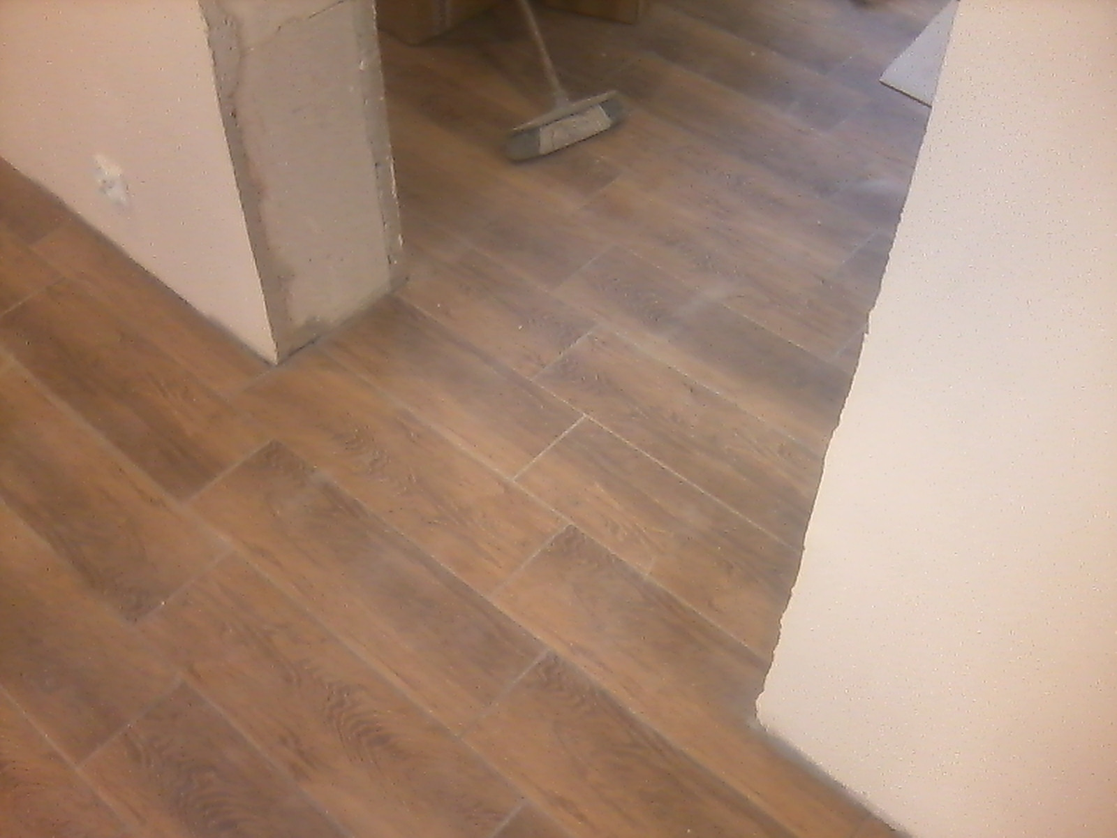 Ułożona pod kątem brązowa podłoga z płytek imitujących drewno, widoczny fragment ściany w trakcie wykończenia i miotła w tle.