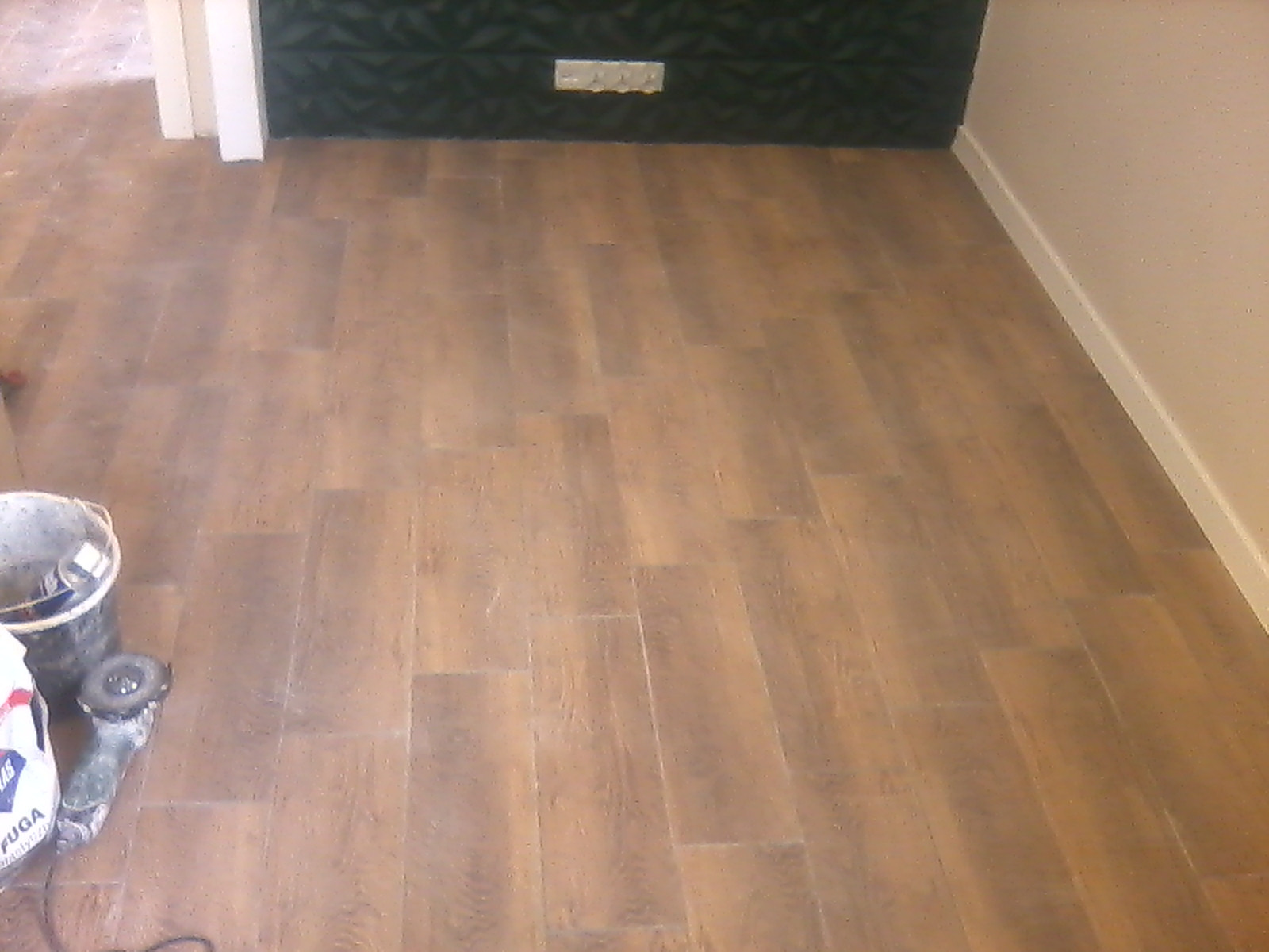 Ułożona podłoga z płytek imitujących drewno, widoczny fragment ściany z dekoracyjną fakturą, wiadro i szlifierka kątowa w lewym dolnym rogu.