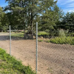 Metalowa siatka ogrodzeniowa z drutem kolczastym na górze, otaczająca teren z drzewami i budynkami w tle, widok z poziomu gruntu.