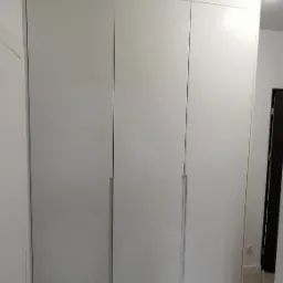 Biała, trzydrzwiowa szafa wnękowa z minimalistycznymi uchwytami, dopasowana do sufitu w jasnym wnętrzu.