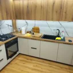 Nowoczesna kuchnia w odcieniach beżu i drewna z blatem imitującym drewno, czarnym zlewem i sprzętem AGD, podświetlona listwą LED.