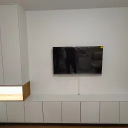 Biała zabudowa meblowa z szafkami wiszącymi i niską komodą pod telewizorem, drewniana wnęka z oświetleniem, minimalistyczny design.