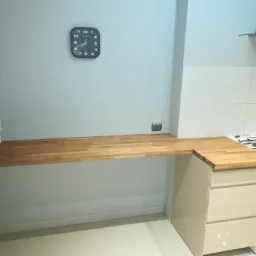 Jasna kuchnia z drewnianym blatem roboczym, szafką z trzema szufladami i kuchenką gazową. Na ścianie wisi czarny zegar.