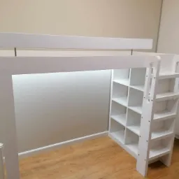 Białe łóżko na antresoli z wbudowanym regałem i drabinką, oświetlone od spodu, na podłodze panele drewnopodobne.