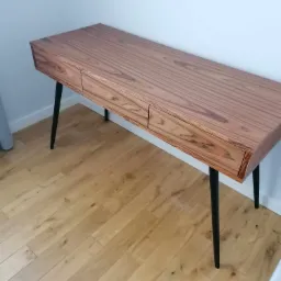 Biurko z drewna egzotycznego o wyraźnym usłojeniu, z dwiema szufladami i czarnymi, skośnymi nogami, na jasnej drewnianej podłodze.