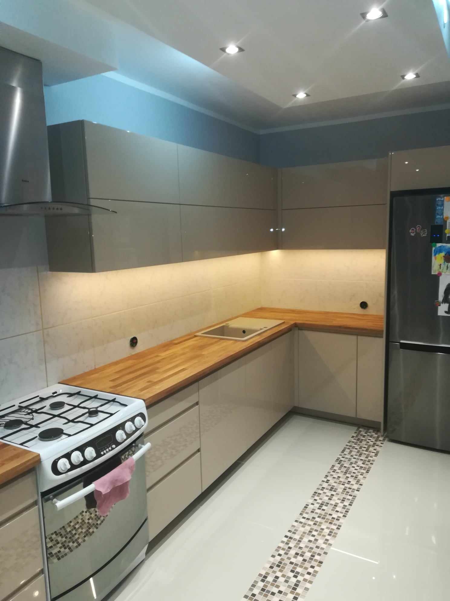 Nowoczesna kuchnia z beżowymi szafkami, drewnianym blatem i białymi płytkami podświetlonymi ledami, z widocznym fragmentem kuchenki z różową ścierką i lodówką.