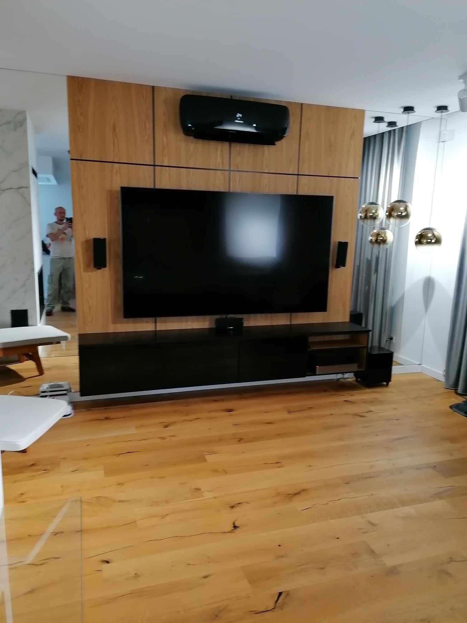 Nowoczesny salon z podłogą z jasnego drewna, ścianą z panelami drewnopodobnymi, na której wisi telewizor i klimatyzator, a pod nim szafka RTV w czarnym połysku. W tle widoczne lampy kuliste...