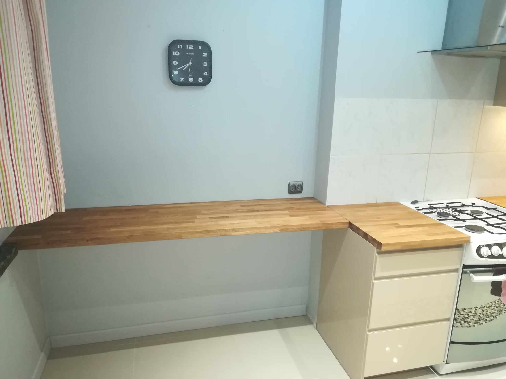 Jasna kuchnia z drewnianym blatem roboczym, szafką z trzema szufladami i kuchenką gazową. Na ścianie wisi czarny zegar.