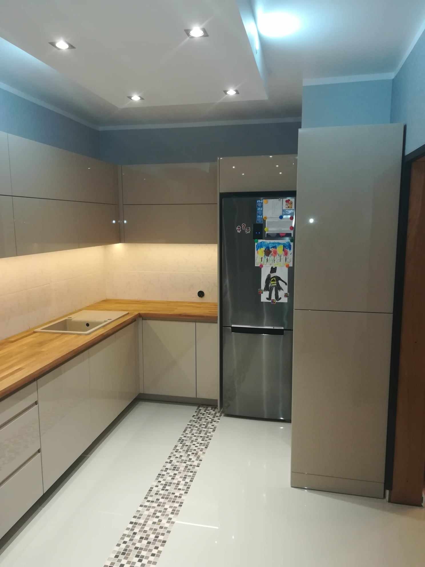 Beżowa zabudowa kuchenna z drewnianym blatem, oświetleniem podszafkowym LED i srebrną lodówką z rysunkami dziecięcymi na białej podłodze z mozaikowym paskiem dekoracyjnym.