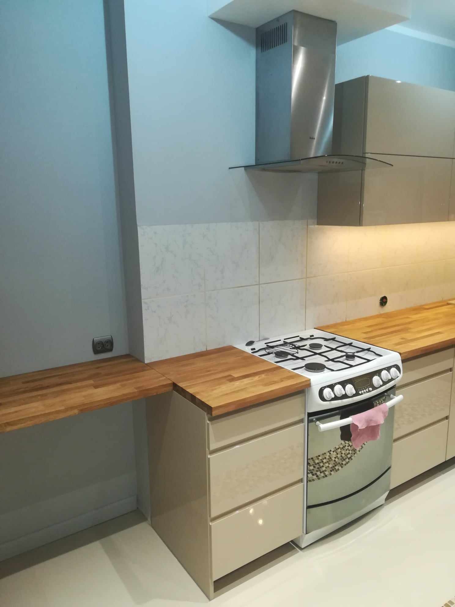 Fragment kuchni z beżowymi szafkami, drewnianym blatem, białą kuchenką gazową i okapem ze stali nierdzewnej. Na piekarniku wisi różowa ścierka.