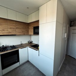 Brat-bud - Nowoczesna kuchnia z drewnianymi i białymi szafkami, czarnym piekarnikiem i płytą grzewczą. Minimalistyczny design, szare płytki podłogowe, drzwi w tle.