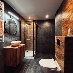 Brat-bud - Nowoczesna łazienka z kabiną prysznicową typu walk-in, umywalką nablatową na drewnianej szafce i podwieszaną toaletą. Ciemne płytki i drewniane akcenty.