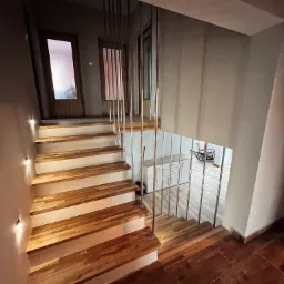 Schody drewniane z metalową balustradą, oświetlenie punktowe na ścianie, widok na pokój z podłogą w jodełkę i bujanym fotelem.