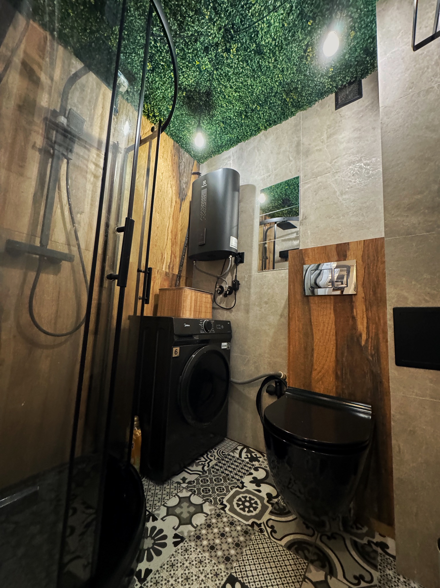 Nowoczesna łazienka z czarną armaturą, kabiną prysznicową, pralką i toaletą. Sufit pokryty sztuczną zielenią. Nietypowe połączenie materiałów i wzorów.