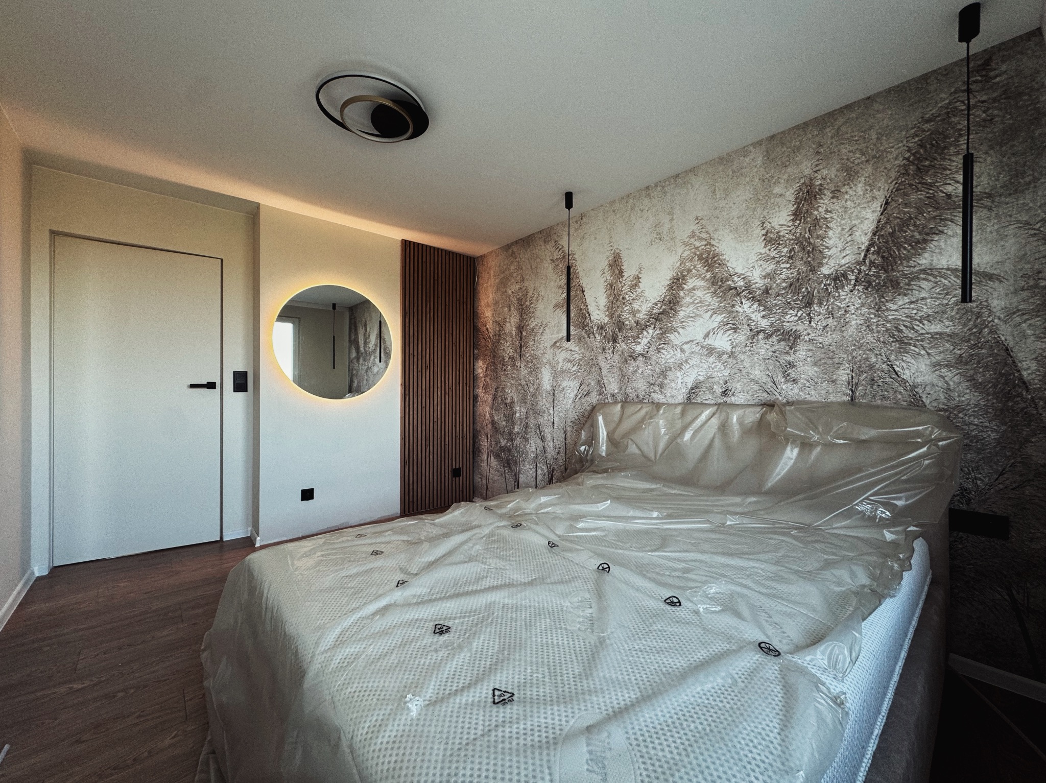 Nowoczesna sypialnia z łóżkiem przykrytym folią ochronną, okrągłe lustro z podświetleniem, dekoracyjna ściana z motywem roślinnym i drewniane panele ścienne.