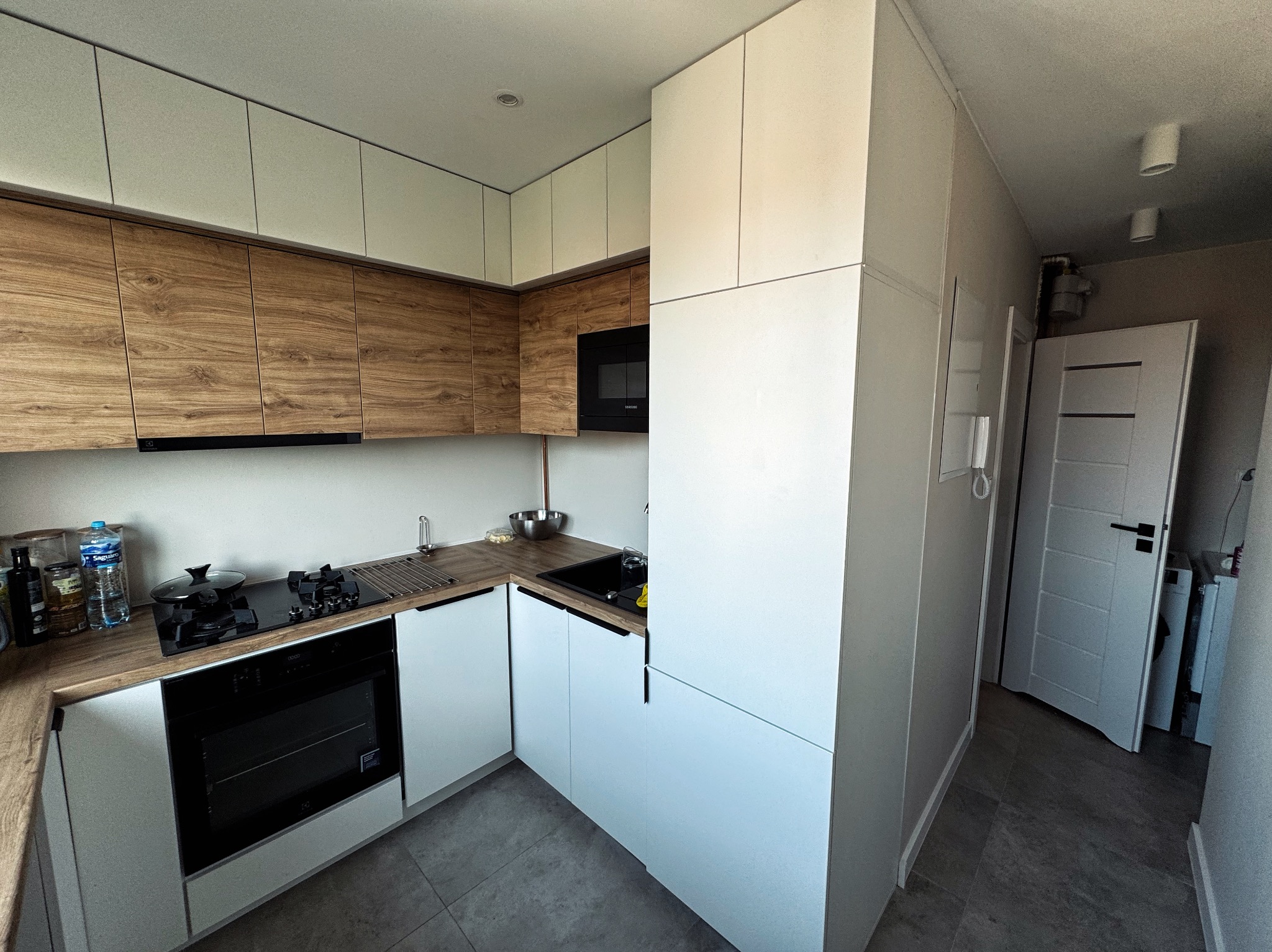 Nowoczesna kuchnia z drewnianymi i białymi szafkami, czarnym piekarnikiem i płytą grzewczą. Minimalistyczny design, szare płytki podłogowe, drzwi w tle.