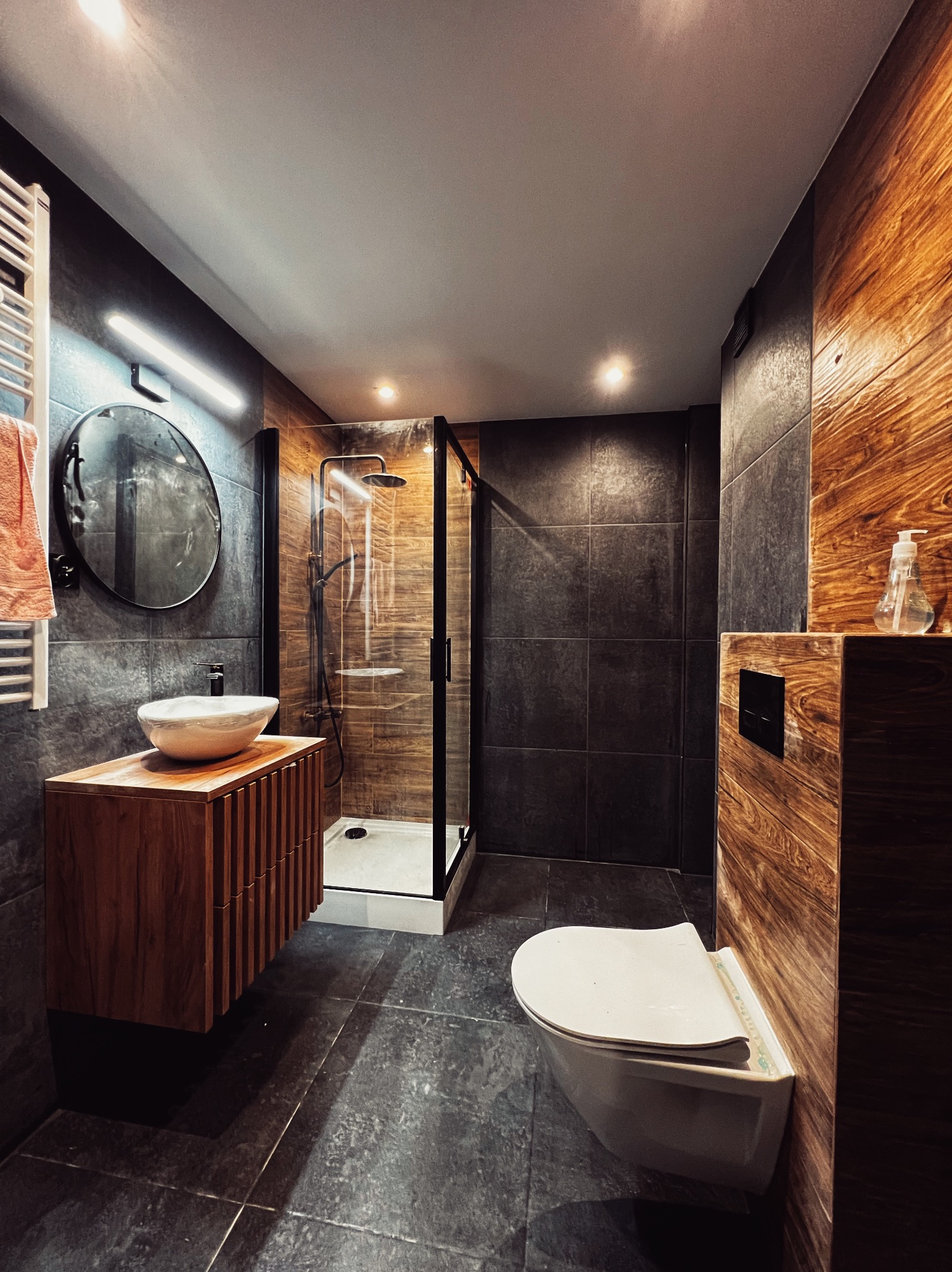 Nowoczesna łazienka z kabiną prysznicową typu walk-in, umywalką nablatową na drewnianej szafce i podwieszaną toaletą. Ciemne płytki i drewniane akcenty.