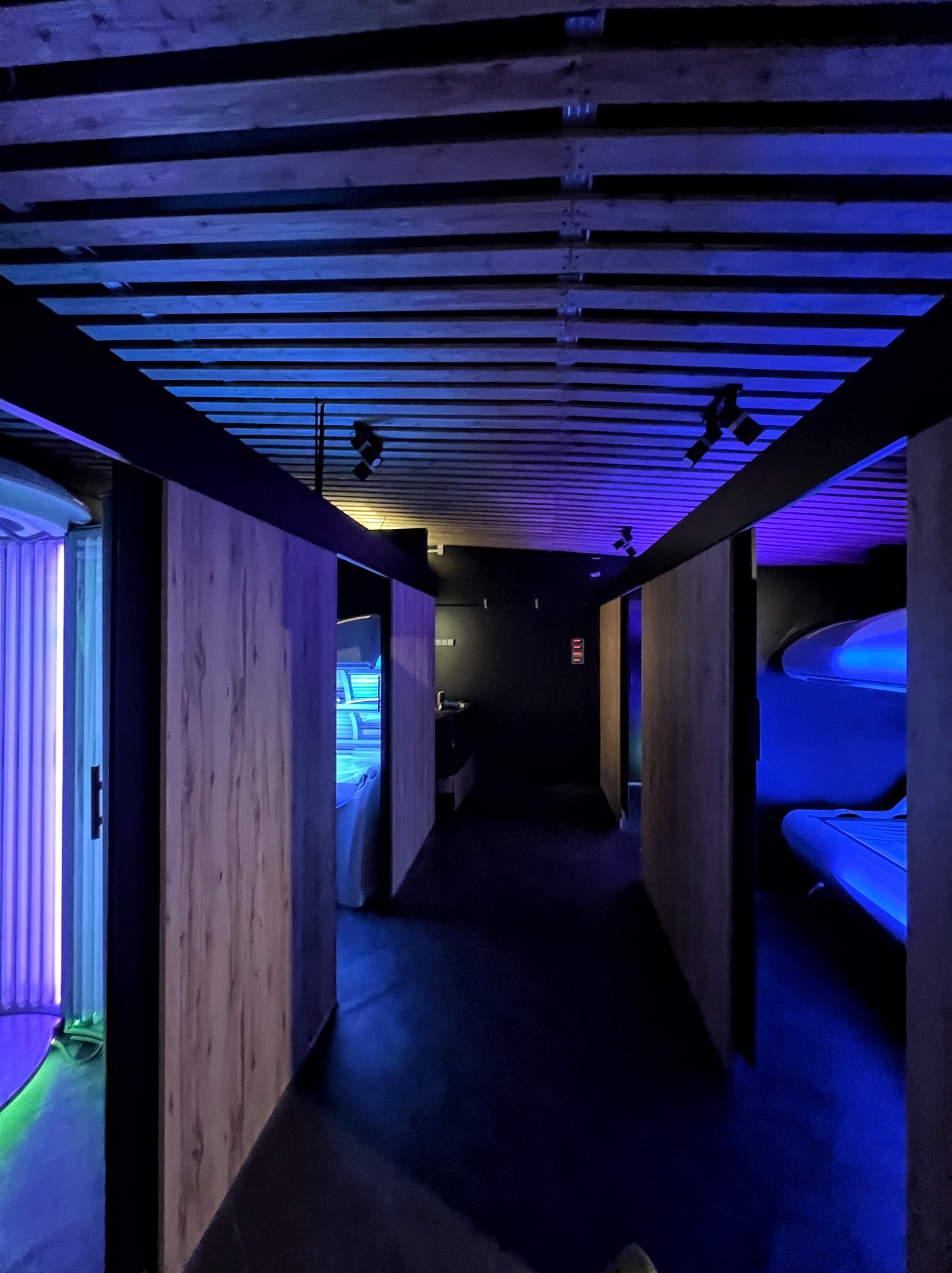 Wnętrze solarium z kabinami opalającymi, oświetlone niebieskim światłem. Widoczne drewniane elementy wykończenia i lampy sufitowe.