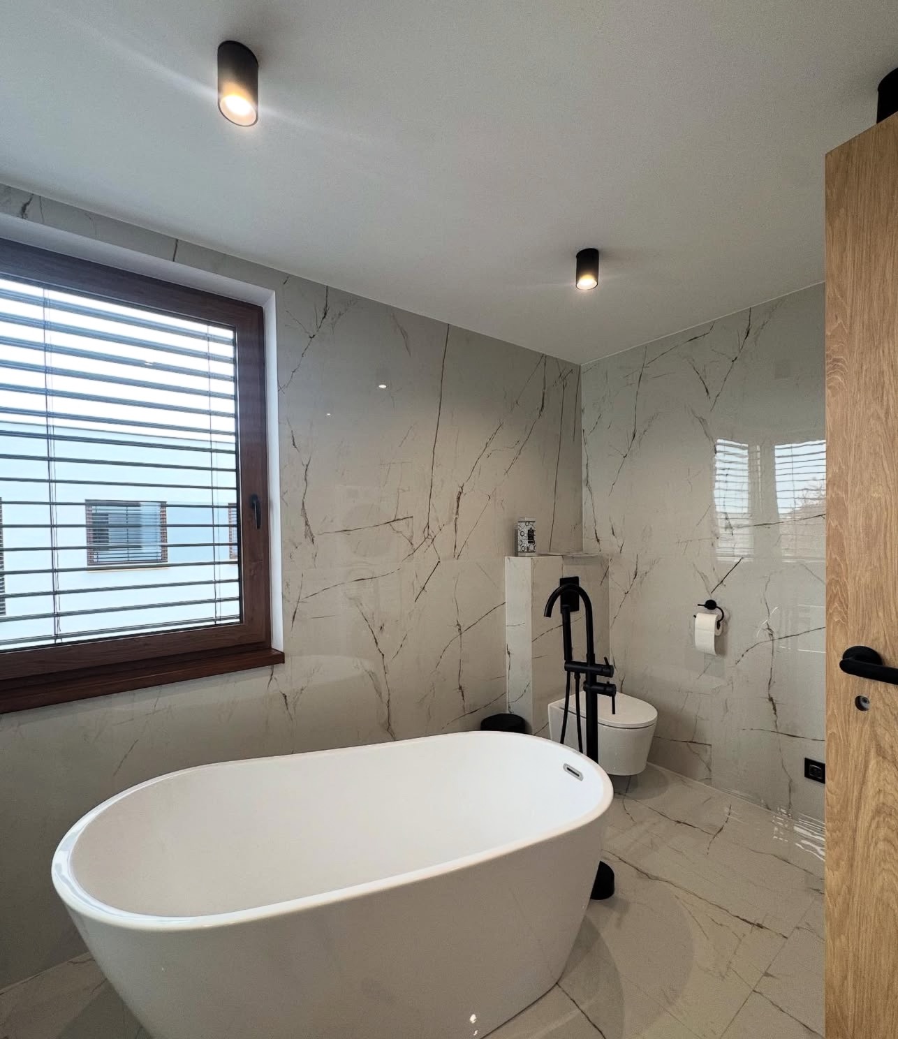 Nowoczesna łazienka z wolnostojącą wanną, marmurowymi płytkami i czarną armaturą. Okno z drewnianą ramą i żaluzjami. Minimalistyczny design.