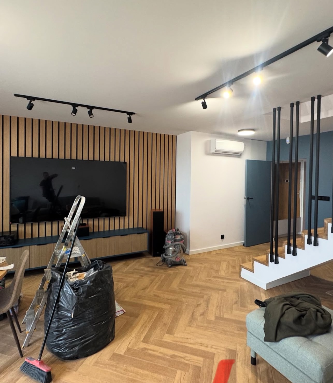 Salon po remoncie z drewnianą ścianą, oświetleniem szynowym i schodami. Widoczny worek na śmieci i drabina, sugerujące prace wykończeniowe.