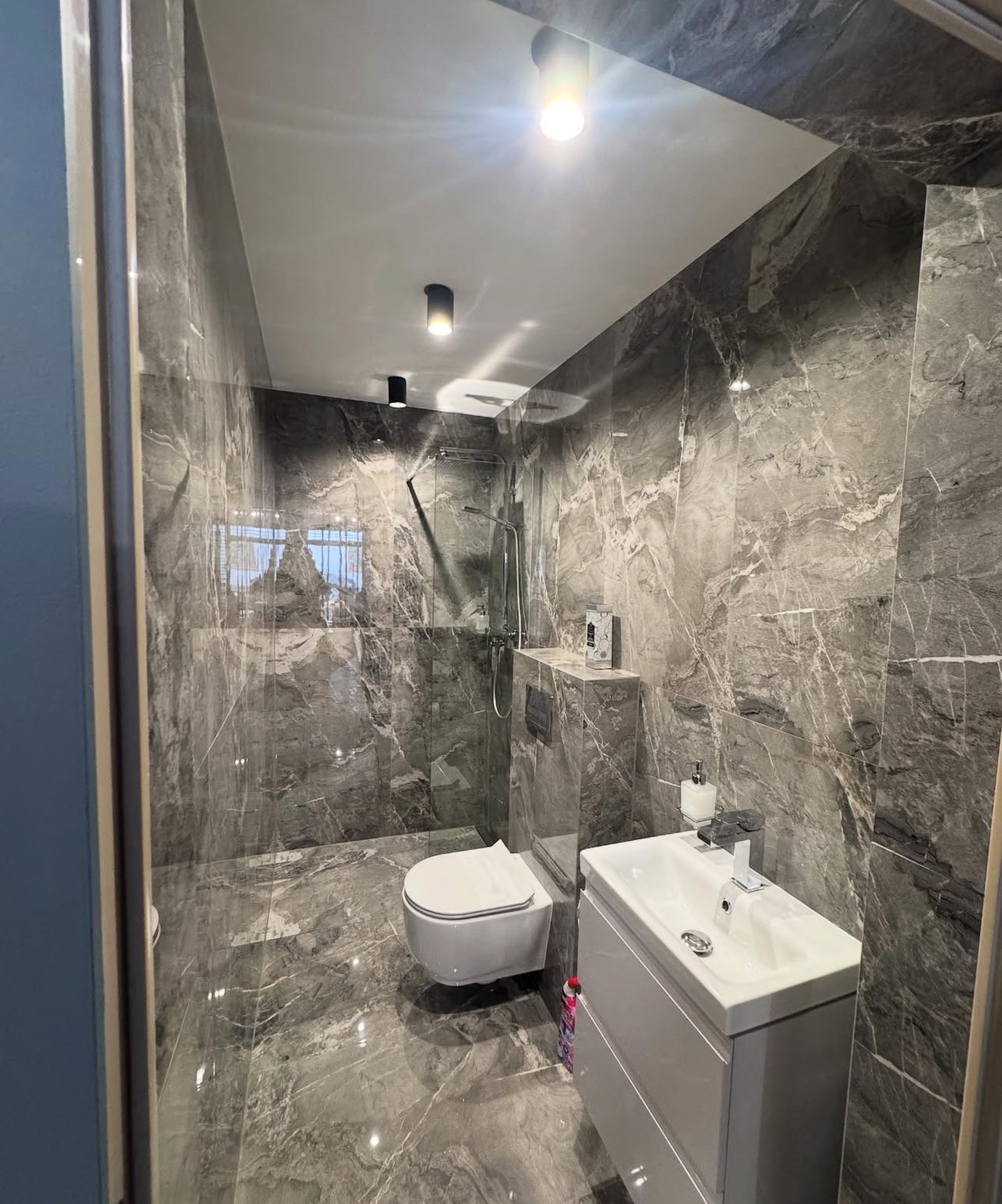 Elegancka łazienka wykończona płytkami imitującymi marmur, z podwieszaną toaletą i nowoczesną umywalką z szafką. Oświetlenie punktowe podkreśla wzór płytek.