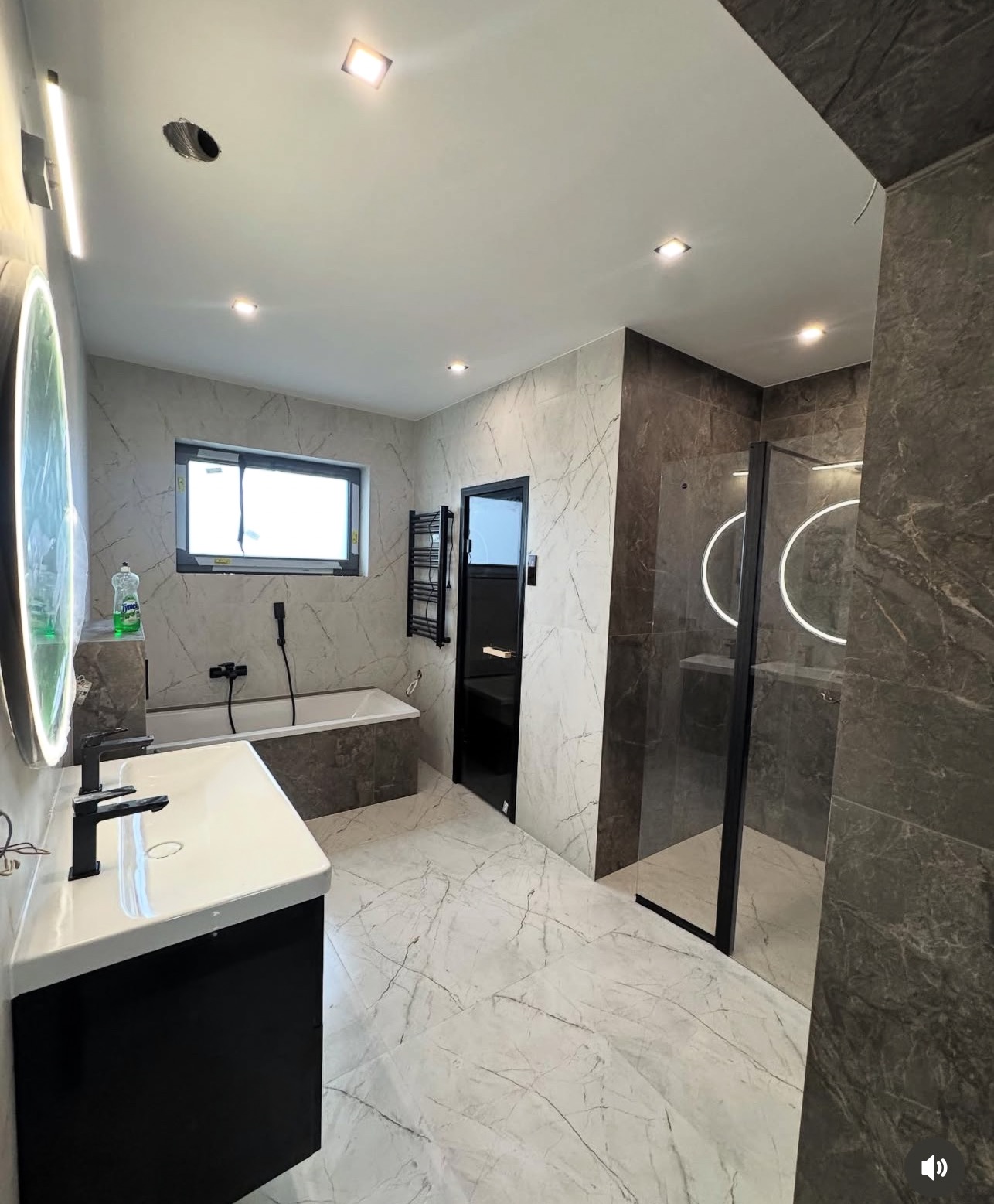 Nowoczesna łazienka z marmurową podłogą i ścianami, czarną armaturą, kabiną prysznicową typu walk-in i podświetlanym lustrem. Minimalistyczny design z akcentami czerni.