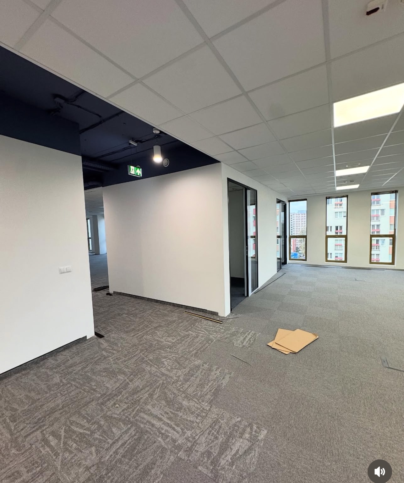 Wnętrze biurowe w trakcie wykańczania: szara wykładzina dywanowa w kwadratach, białe ściany, sufit kasetonowy, okna z widokiem na budynki. Kartony na podłodze.