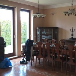 MIKO Perfekt - Duży salon z brązowymi płytkami na podłodze, drewnianym stołem i krzesłami, oraz oknami z widokiem na zieleń