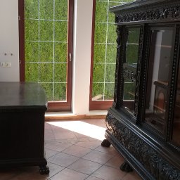 MIKO Perfekt - Wnętrze pokoju z antycznymi meblami: ciemna, rzeźbiona komoda i witryna na tle okien z widokiem na zielony żywopłot, terakotowa podłoga w kwadraty, oświetlenie sufitowe z kloszem.