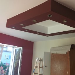 MIKO Perfekt - Pomieszczenie w trakcie remontu z pomalowaną na bordowo ścianą i sufitem podwieszanym z oświetleniem halogenowym, widoczna drabina i okno z widokiem na zewnątrz.