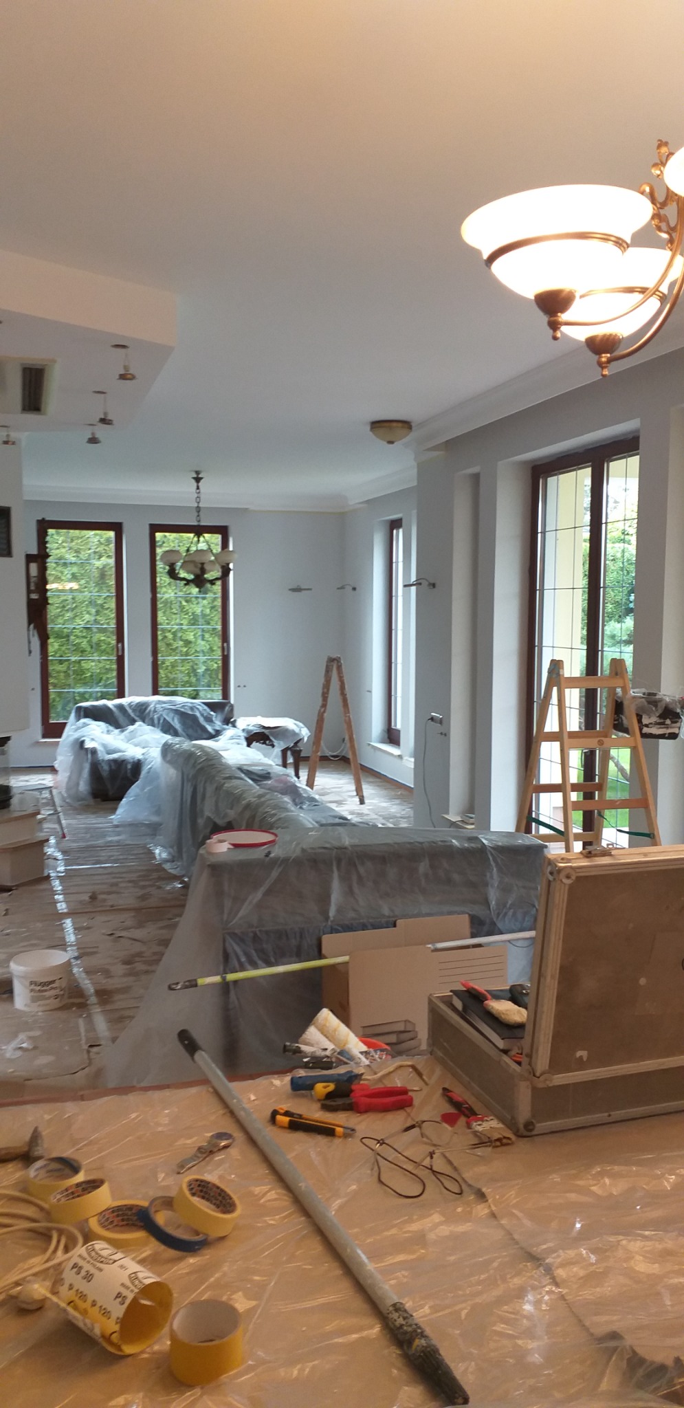 Przygotowania do malowania wnętrza: meble przykryte folią ochronną, widoczne drabiny, pędzle, wałki malarskie, taśmy malarskie i skrzynka z narzędziami na podłodze zabezpieczonej folią.