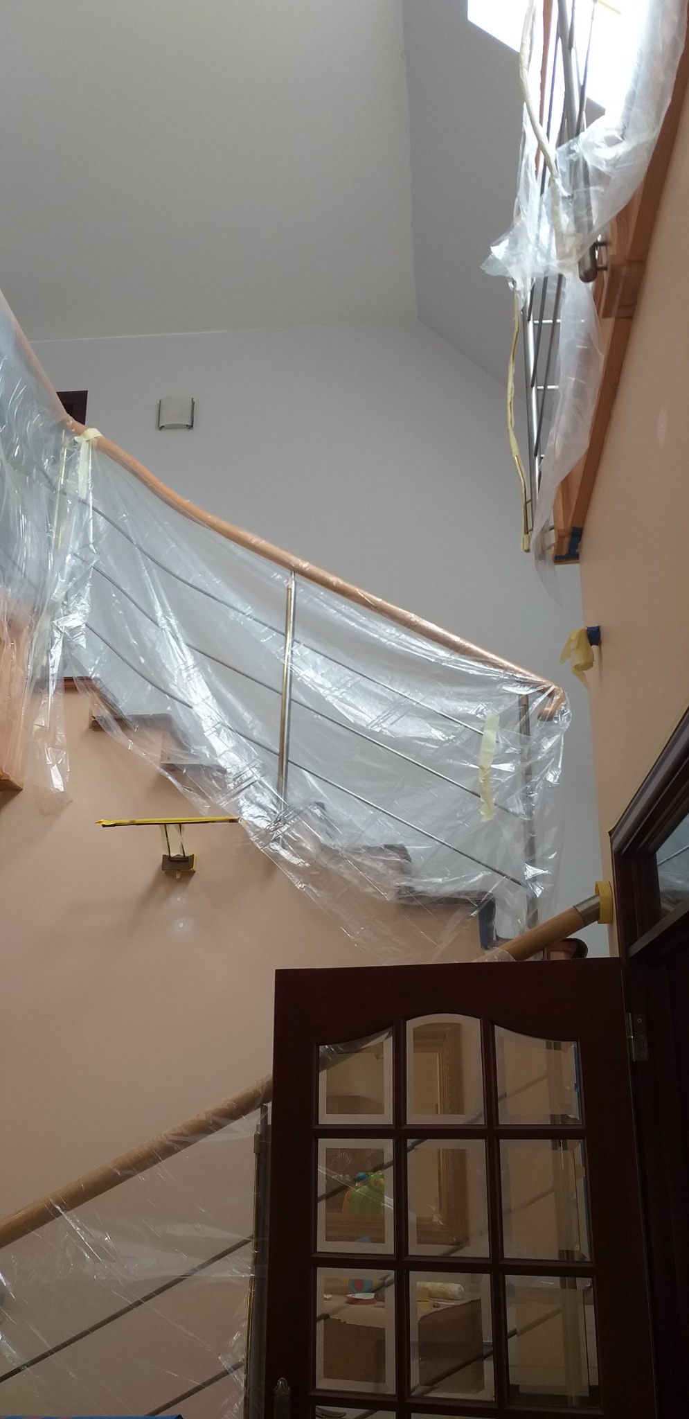 Schody z metalową balustradą zabezpieczone folią malarską przed rozpoczęciem prac, widoczna brązowa ościeżnica drzwiowa z szybami.