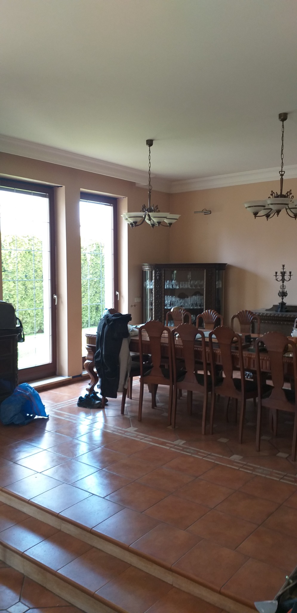 Duży salon z brązowymi płytkami na podłodze, drewnianym stołem i krzesłami, oraz oknami z widokiem na zieleń