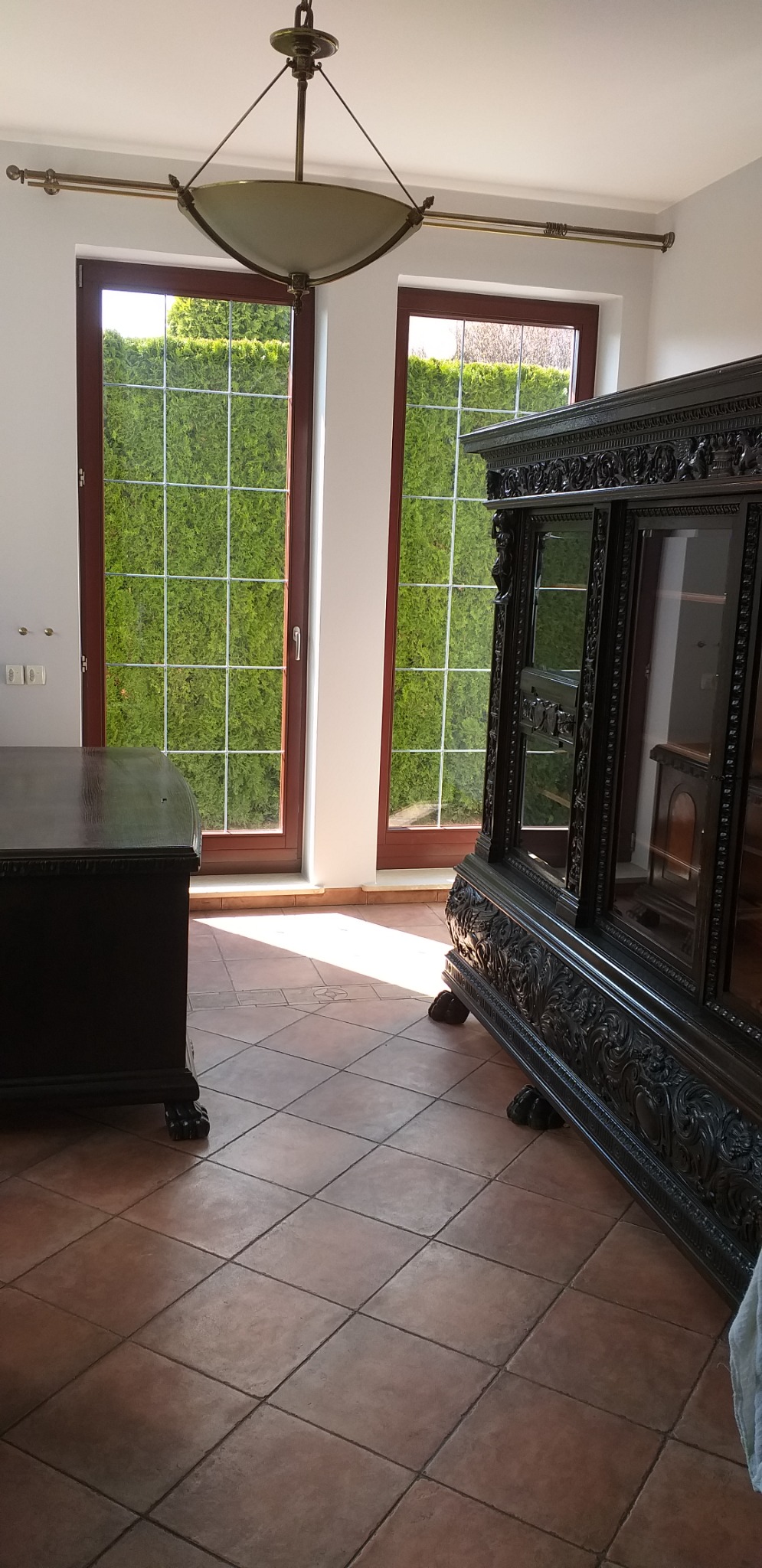 Wnętrze pokoju z antycznymi meblami: ciemna, rzeźbiona komoda i witryna na tle okien z widokiem na zielony żywopłot, terakotowa podłoga w kwadraty, oświetlenie sufitowe z kloszem.