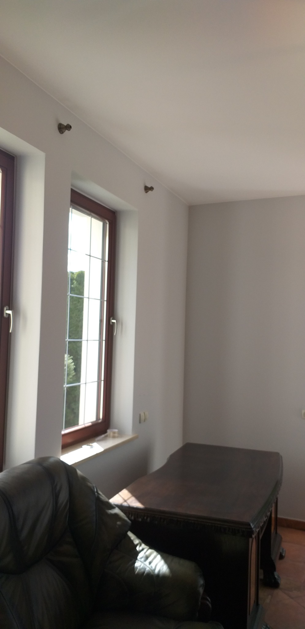 Jasne wnętrze pokoju z dwoma oknami ze szprosami, pomalowanymi na brązowo, z widokiem na zieleń na zewnątrz, świeżo pomalowane ściany na szaro, widoczne uchwyty karniszy pod sufitem, duży, ciemny...